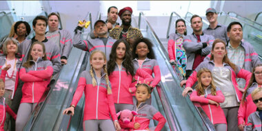 Elenco de Feel the Beat na escada rolante. São três escadas rolantes, uma do lado da outra. Na foto há crianças e adultos, todos vestindo o uniforme rosa e cinza da escola de dança.