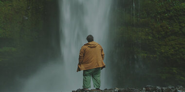 O ator Jorge Garcia parado em frente à uma cachoeira, numa montanha pedregosa. Ele está de costas para o espectador, trajando um casaco amarelo e uma calça jeans desgastada, meio verde. Ao redor da cachoeira há vários matos verdes.